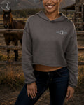 Woman wearing a gray hoodie with a logo, standing in a rustic outdoor setting with a horse in the background.